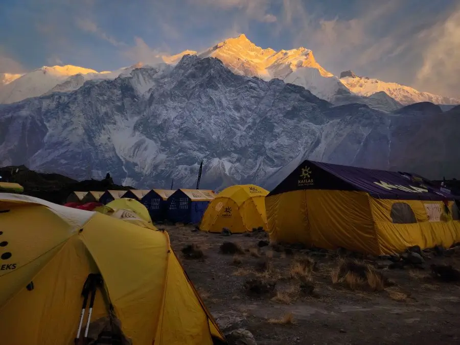 A cluster of yellow, blue, and purple tents forms the Annapurna base camp, set against a rocky terrain. The majestic Annapurna I, snow-covered and glowing under the sunset, towers in the background under a sky with scattered clouds.