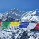 Colorful Tibetan prayer flags flutter in the wind against a backdrop of snow-covered Himalayan peaks, with Annapurna’s majestic, icy ridgelines rising under a clear blue sky.