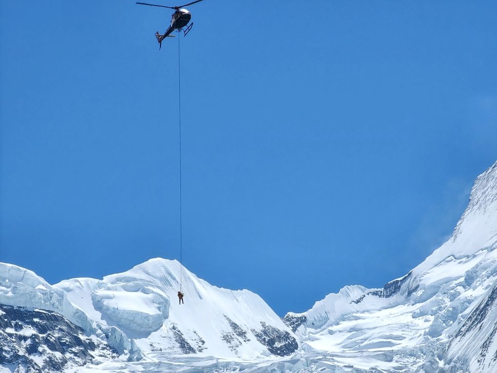 A high-altitude rescue operation on Annapurna: a helicopter hovers above the snowy peaks, airlifting a climber suspended by a long rope beneath it.