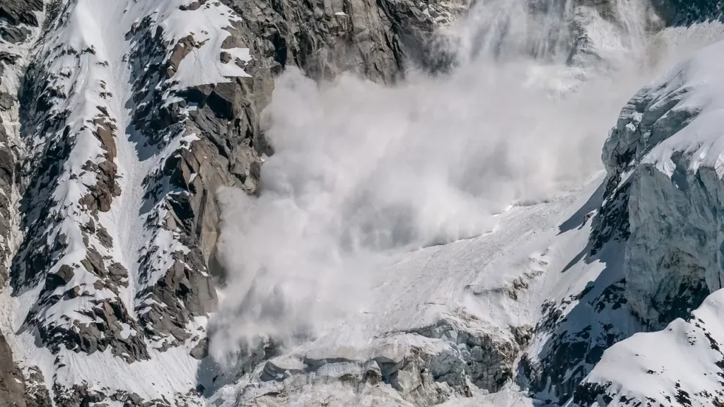 A massive avalanche cascades down a steep, snow-covered mountain, sending a cloud of snow billowing outward. The rugged peaks on either side are dusted with snow, with rocky outcrops protruding through the icy slopes. The scene captures the raw power of nature in a remote alpine environment.