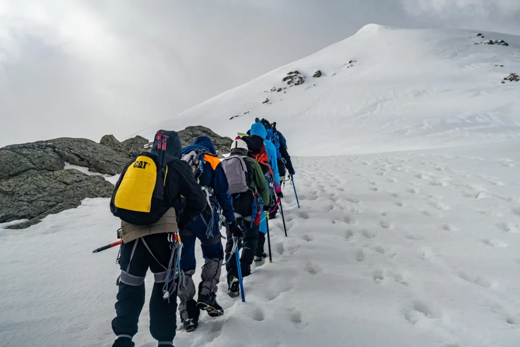 A group of mountaineers, dressed in colorful jackets and equipped with backpacks, ascends a snowy mountain slope. They use trekking poles and are roped together, leaving a trail of footprints in the snow. The landscape features rocky outcrops and a snow-covered peak under a cloudy sky, showcasing the challenging terrain of their climb.