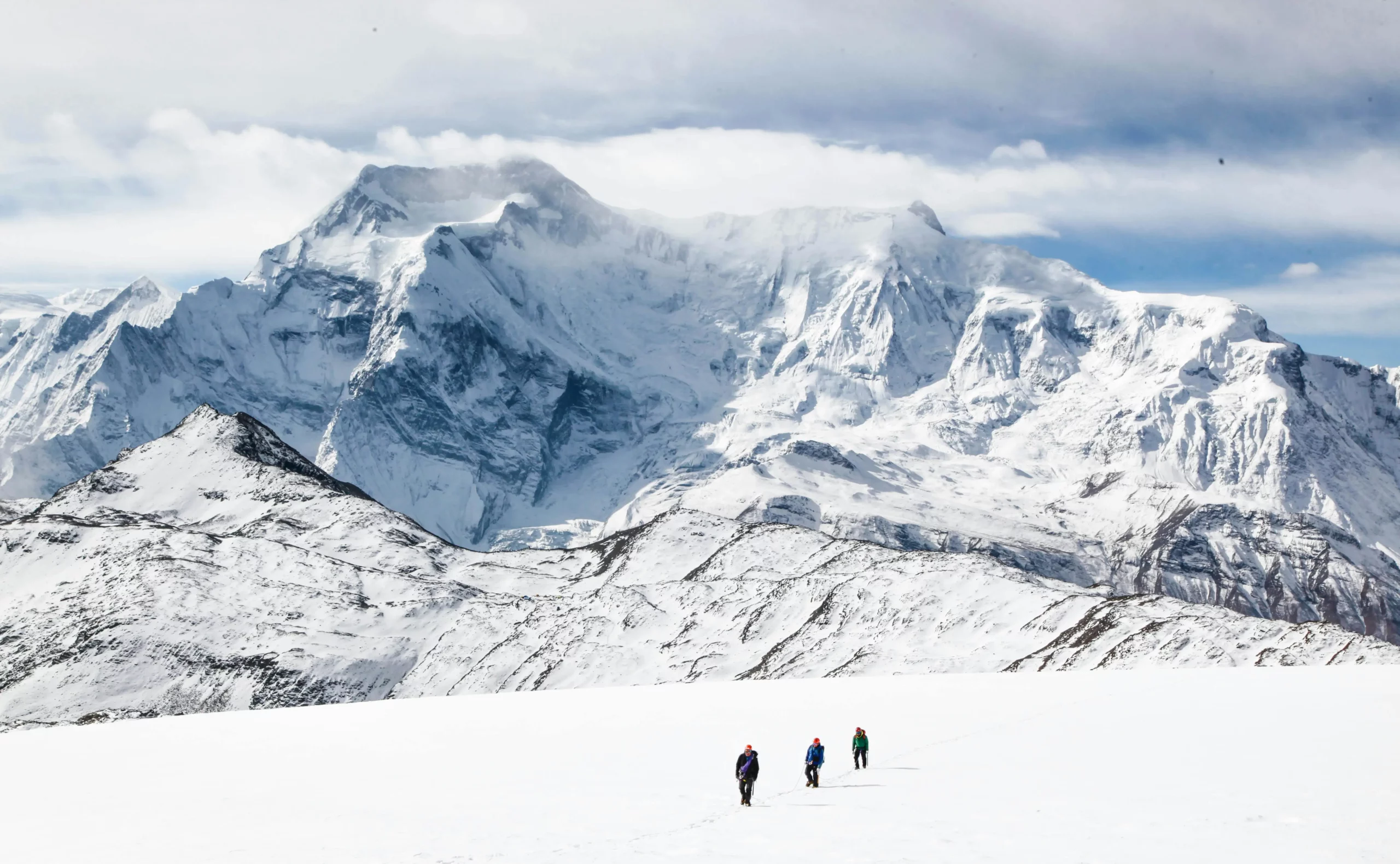 Three climbers in colorful jackets cross a snowy expanse, with towering, snow-covered Himalayan mountains under a partly cloudy sky.