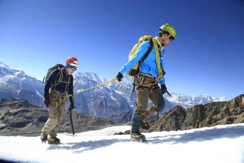 Two alpine climbers traverse a snowy mountain slope, roped together for safety. They wear helmets, crampons, and backpacks, with ice axes in hand, against a backdrop of rugged peaks and a clear blue sky.