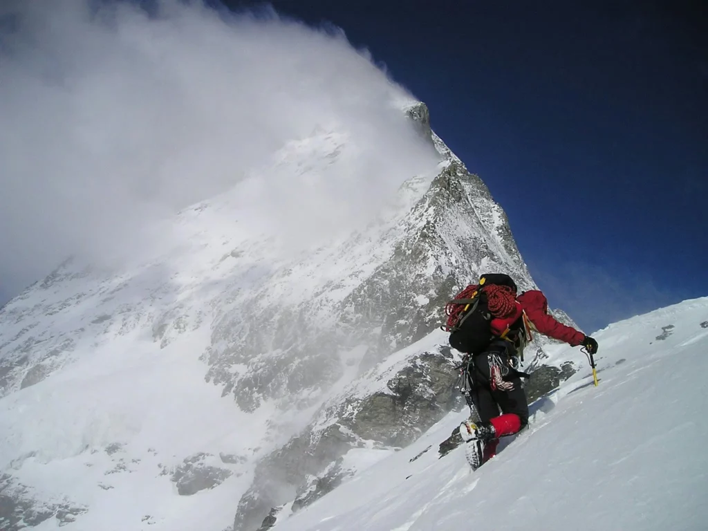 A mountaineer in a red jacket and black gear ascends a steep, snow-covered mountain slope. They carry a black backpack with red climbing ropes coiled on top and use an ice axe for support. The towering peak ahead is shrouded in swirling clouds, with a clear blue sky contrasting the snowy, rocky terrain. The scene captures the intensity of high-altitude climbing.