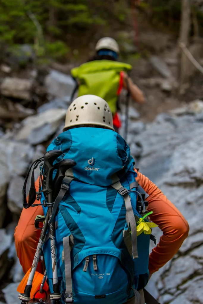 Two mountaineers climb a rocky mountain path surrounded by trees. The lead climber wears a white helmet, orange jacket, and a blue Deuter backpack with trekking poles attached, featuring a yellow flower tucked into a strap. Behind them, another climber in a green jacket and white helmet navigates the steep terrain, with blurred rocks and greenery in the background.