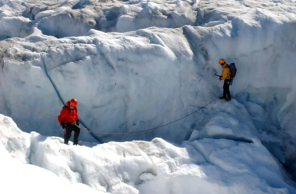 Two mountaineers navigate a crevasse-filled glacier, roped together with a red climbing rope. The lead climber, in a red jacket and helmet, carefully steps across the icy terrain, while the second, in a yellow jacket and orange helmet, holds an ice axe and follows closely. Towering ice walls and deep snow surround them in a remote alpine setting.