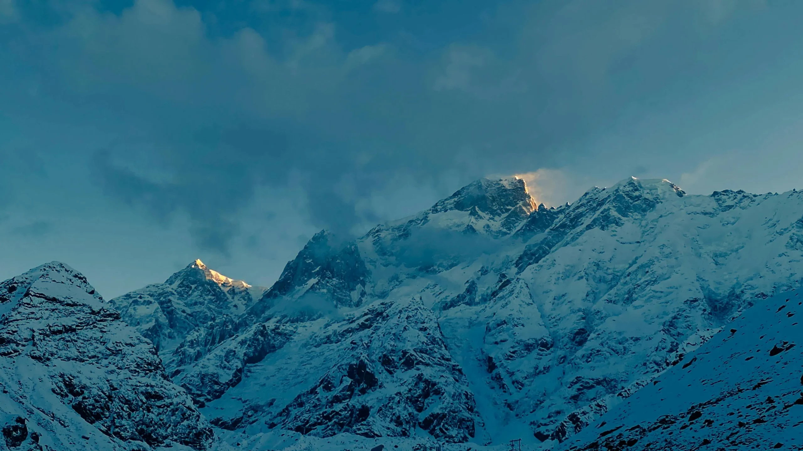 Snow-covered mountain peaks bathed in soft golden sunlight under a moody, cloud-filled sky.