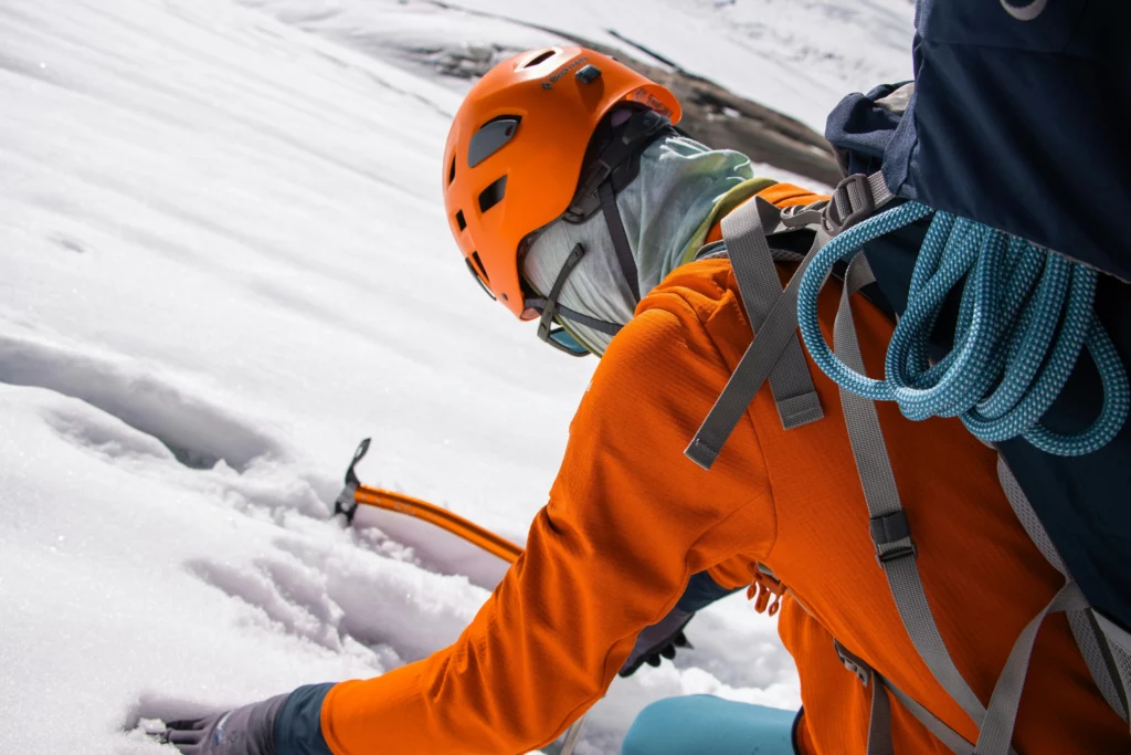 A mountaineer in an orange helmet and jacket climbs a snowy slope, using an ice axe to dig into the snow. They wear a large blue backpack with a coiled teal climbing rope attached to the side. The background features a snow-covered landscape with a glimpse of distant mountains under a cloudy sky.
