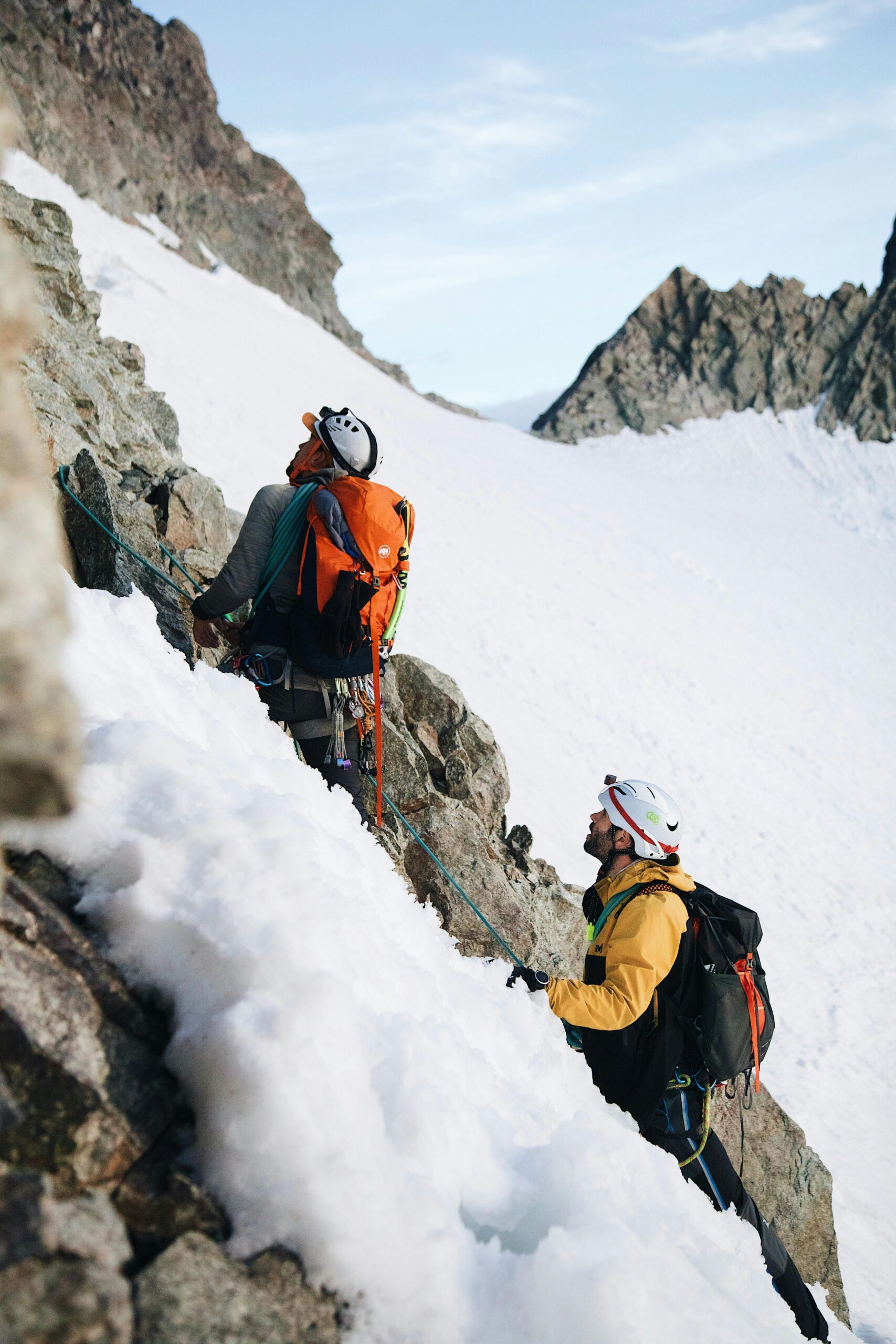 Two mountaineers climb a steep, snowy, and rocky mountain face, roped together with a teal climbing rope. The lead climber, in a white helmet and gray jacket, carries an orange backpack, while the second climber wears a yellow jacket and white helmet with a black backpack. The background features snow-covered slopes and rugged peaks under a light blue sky with scattered clouds.