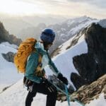 A mountaineer in a blue helmet and teal jacket climbs a snowy, rocky slope in a mountainous landscape. She carries a large yellow backpack and are secured with a teal climbing rope. The background features snow-covered peaks and a vast, misty valley under a partly cloudy sky, with soft sunlight illuminating the scene.