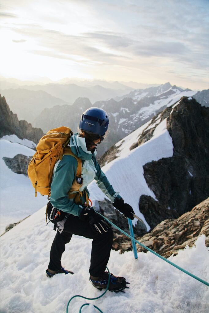 A mountaineer in a blue helmet and teal jacket climbs a snowy, rocky slope in a mountainous landscape. She carries a large yellow backpack and are secured with a teal climbing rope. The background features snow-covered peaks and a vast, misty valley under a partly cloudy sky, with soft sunlight illuminating the scene.