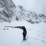 A long line of climbers treks through deep snow toward Everest’s Camp I, surrounded by towering ice cliffs and jagged mountains, under an overcast sky with tents visible in the background.