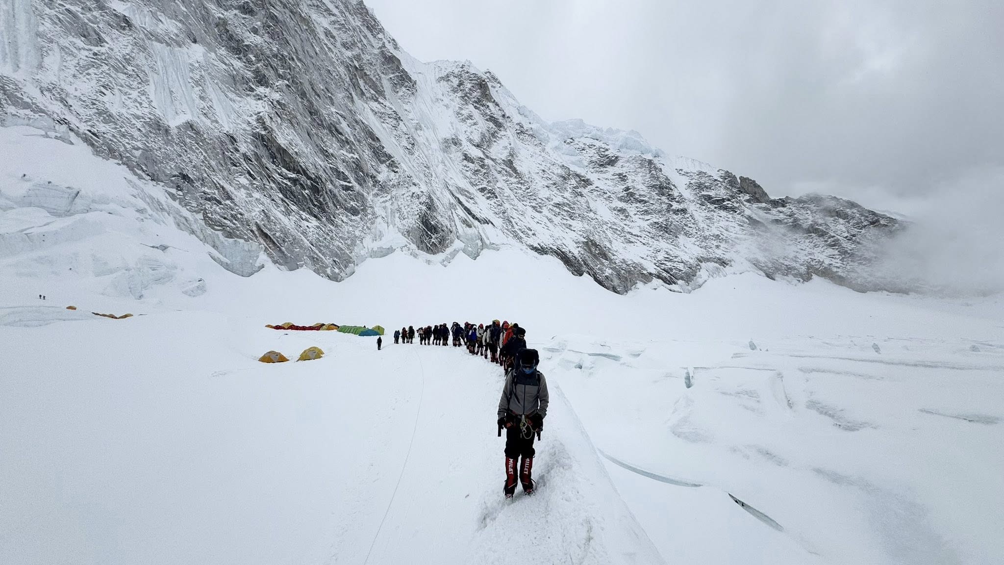A long line of climbers treks through deep snow toward Everest’s Camp I, surrounded by towering ice cliffs and jagged mountains, under an overcast sky with tents visible in the background.