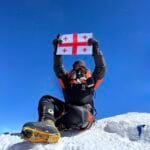 A climber wearing full mountaineering gear and an oxygen mask sits on the snowy summit of Mount Everest, holding the Georgian flag above their head. The bright blue sky and snow-covered peak emphasize the high altitude and extreme conditions.