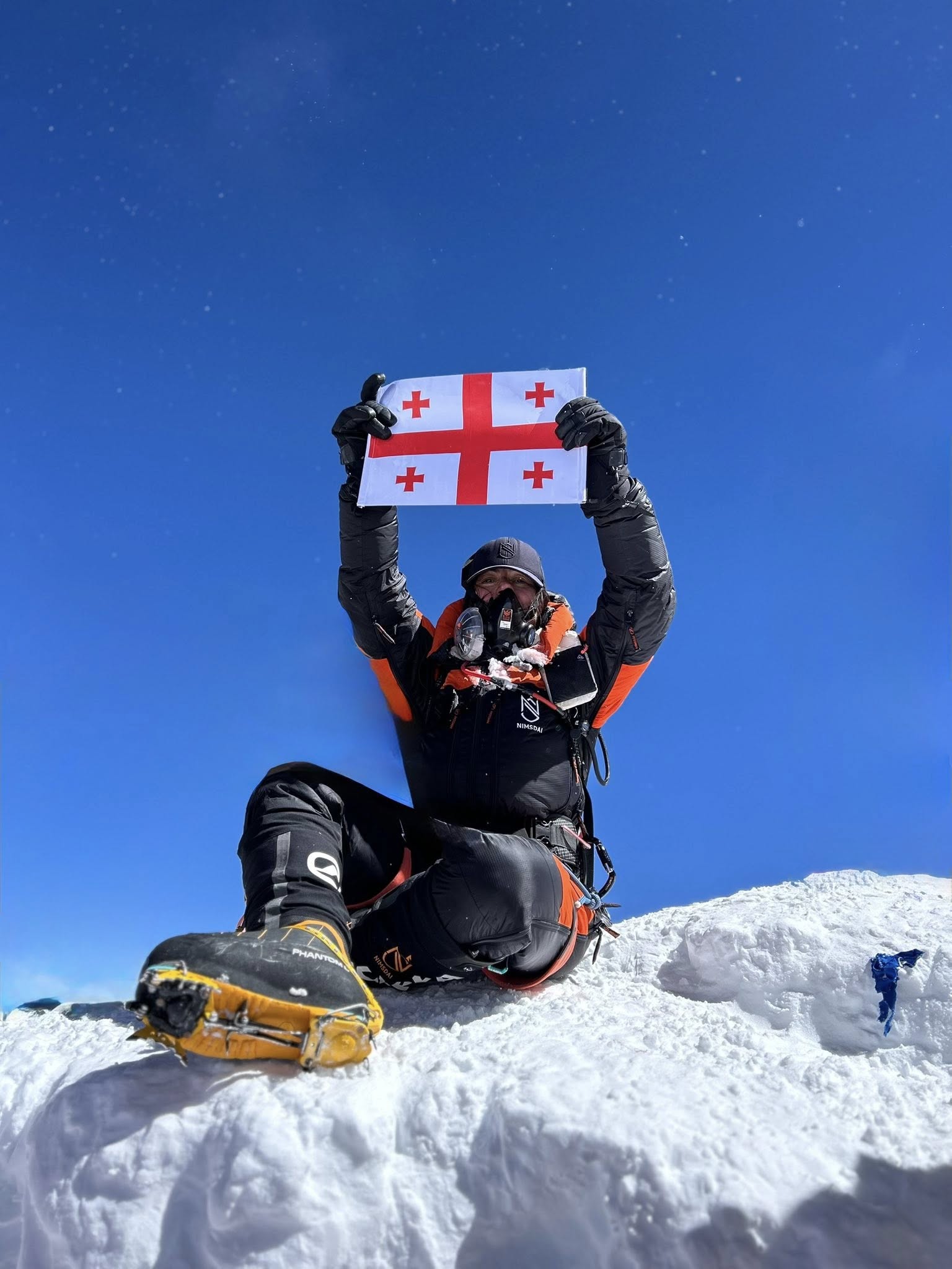 A climber wearing full mountaineering gear and an oxygen mask sits on the snowy summit of Mount Everest, holding the Georgian flag above their head. The bright blue sky and snow-covered peak emphasize the high altitude and extreme conditions.