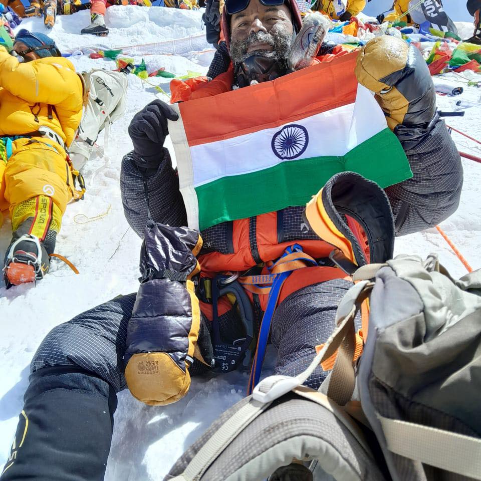 Navdeep Sood, wearing a down suit and oxygen mask, sits on the snow at the summit of Mount Everest holding the Indian national flag. Surrounded by other climbers in brightly colored gear, he appears visibly exhausted but triumphant, with frost on his face and a background of prayer flags and snow-covered terrain.