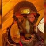 A person inside a brightly lit orange tent is wearing a black Summit Oxygen full-face oxygen mask with side filters. They are also wearing a headlamp labeled "Black Diamond" over a dark beanie. Only the upper part of their face is visible through the mask.
