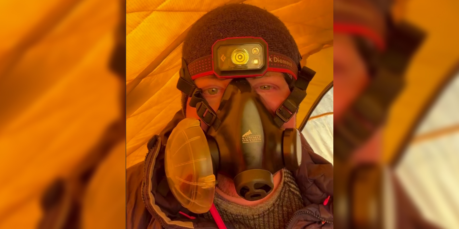 A person inside a brightly lit orange tent is wearing a black Summit Oxygen full-face oxygen mask with side filters. They are also wearing a headlamp labeled "Black Diamond" over a dark beanie. Only the upper part of their face is visible through the mask.