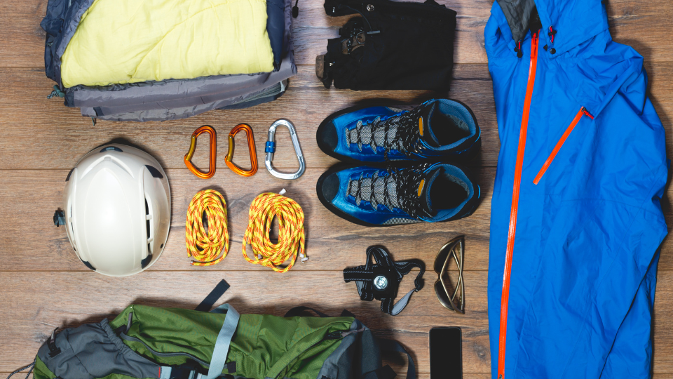 A flat lay of mountaineering gear arranged on a wooden floor, including a sleeping bag, helmet, climbing rope, carabiners, hiking boots, gloves, blue waterproof jacket, backpack, sunglasses, compass, and a smartphone.