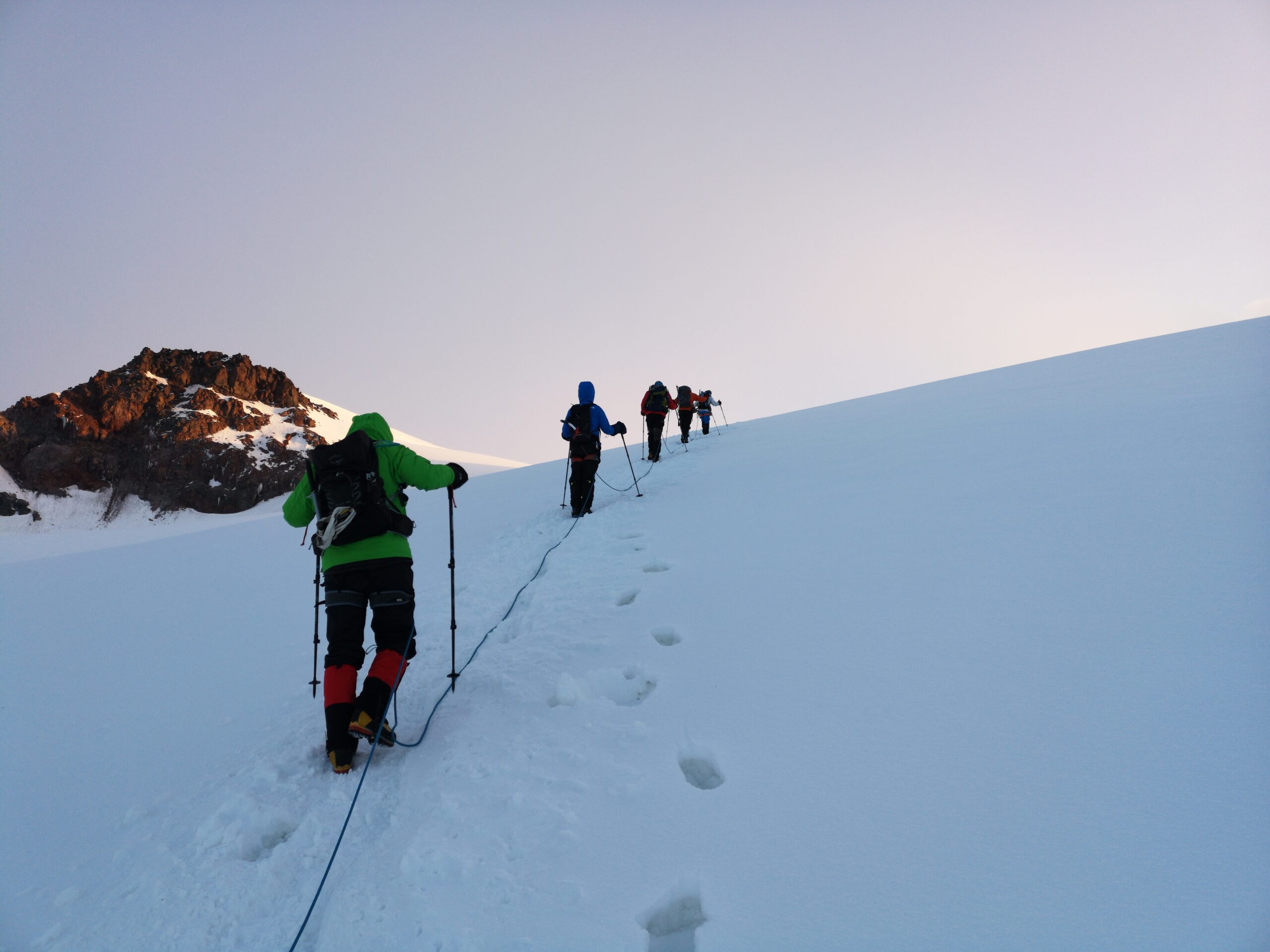 A group of climbers in colorful jackets treks across a snow-covered slope on Mount Kazbek, with its rugged, icy peak rising sharply in the background. The partly cloudy sky casts a soft light over the dramatic, glacier-strewn landscape of the Caucasus Mountains.