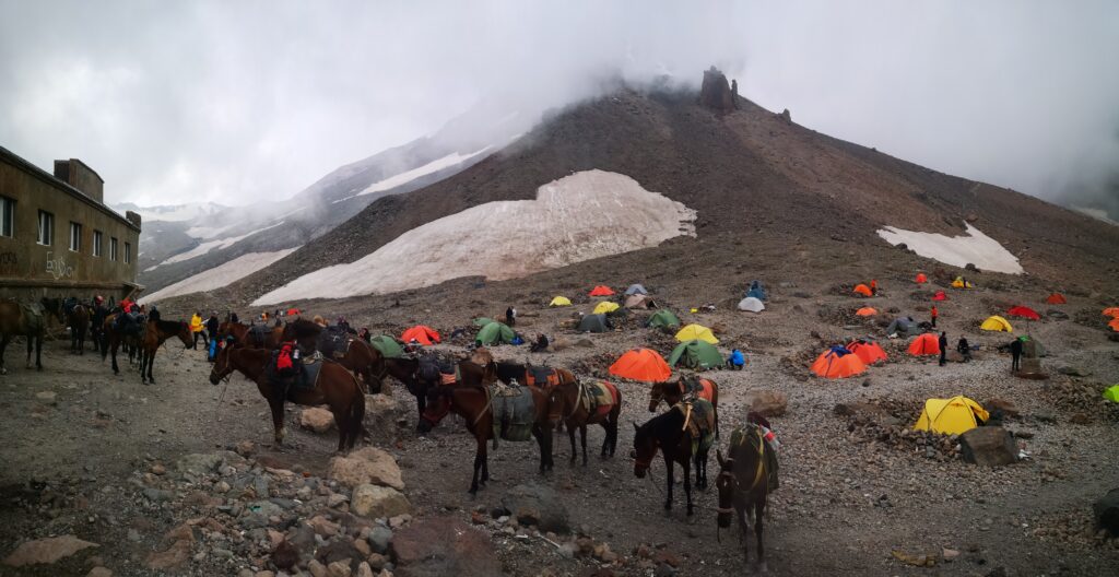Colorful mountaineering tents scattered across a rocky alpine base camp at the foot of Mount Kazbek, Georgia, with steep ridges and a snow-covered peak rising in the background under a blue sky. Climbers can be seen preparing gear amid stone windbreaks and glacial terrain.
