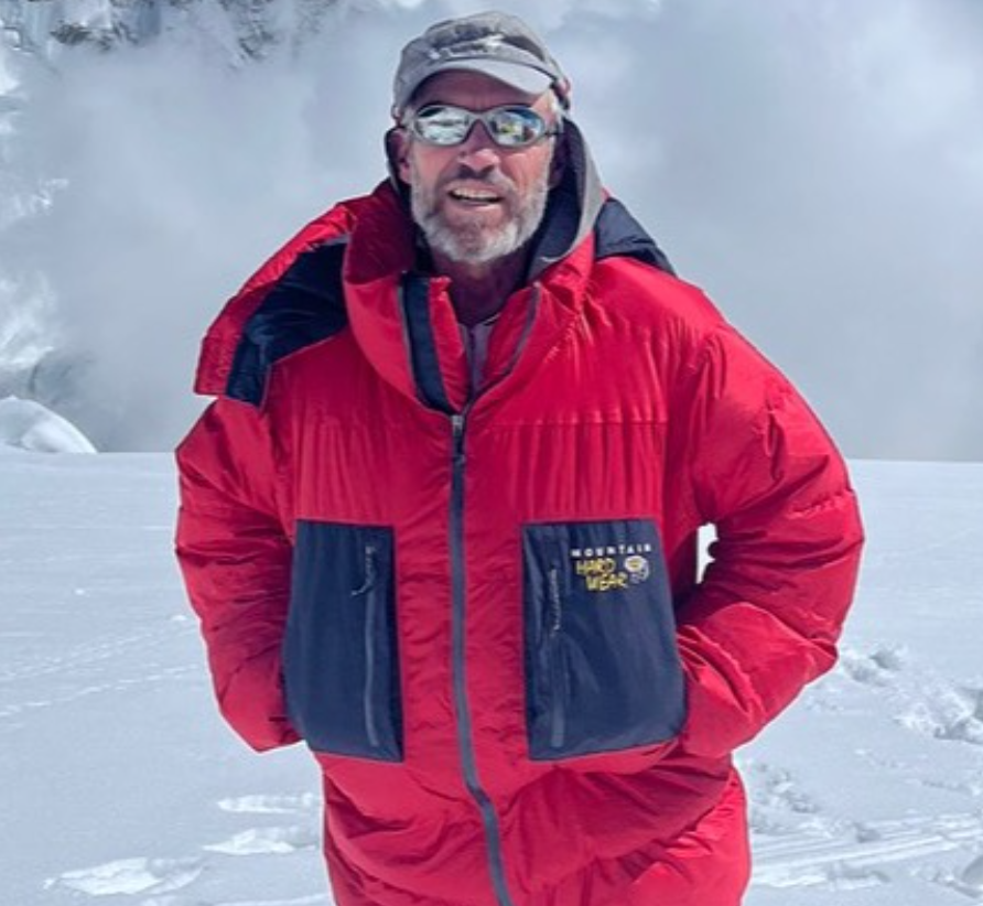 A man wearing a red and navy Mountain Hardwear expedition parka stands on a snowy mountain slope, with snow and ice formations in the background. He has a gray cap, reflective sunglasses, and a light beard, appearing prepared for high-altitude conditions.