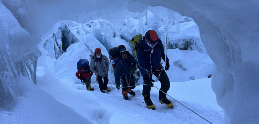 A group of climbers, equipped with helmets, backpacks, and climbing gear, navigates through a narrow, icy crevasse in a glacier. They are roped together for safety, with snow and ice walls towering around them, creating a dramatic and cold mountainous environment.