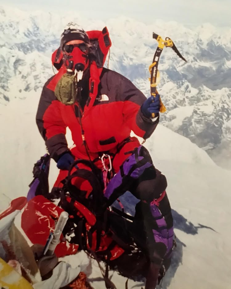 **Alt text:**
A mountaineer in a red and black down suit sits on a snowy summit, wearing an oxygen mask and holding an ice axe in one hand. Surrounded by snow-covered peaks under a clear sky, the climber appears to be at a high-altitude Himalayan summit, possibly Mount Everest. Mountaineering gear is visible at their feet, including ropes and a backpack.

