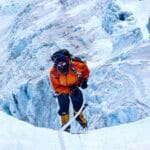 A high-altitude mountaineer wearing an orange jacket, black pants, and yellow climbing boots ascends a steep, icy slope using a rope. The background reveals deep crevasses and rugged ice formations, indicating a dangerous and remote glacier environment, likely on a Himalayan peak.