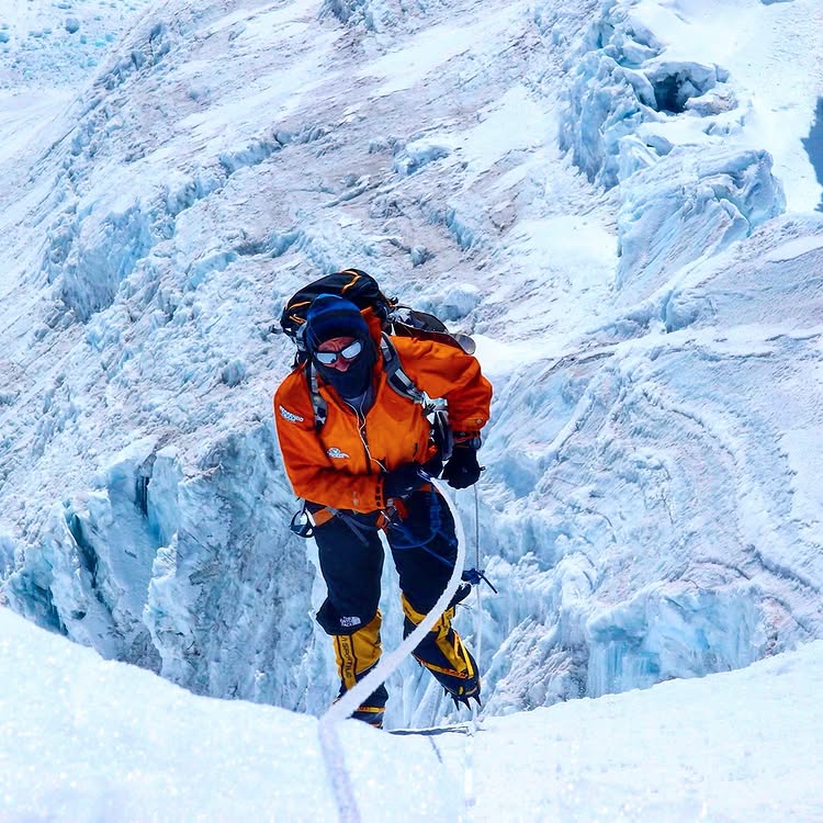 A high-altitude mountaineer wearing an orange jacket, black pants, and yellow climbing boots ascends a steep, icy slope using a rope. The background reveals deep crevasses and rugged ice formations, indicating a dangerous and remote glacier environment, likely on a Himalayan peak.