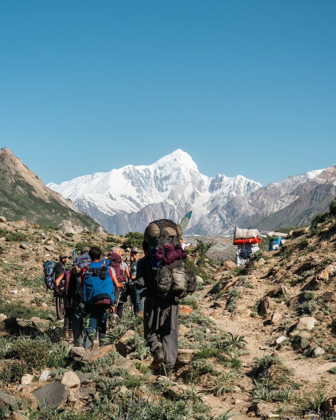 A group of hikers with backpacks walks along a rocky trail in a mountainous region, with a snow-capped peak dominating the background under a clear blue sky. Some porters carry large loads on their backs, and the landscape features sparse vegetation and rugged terrain.
