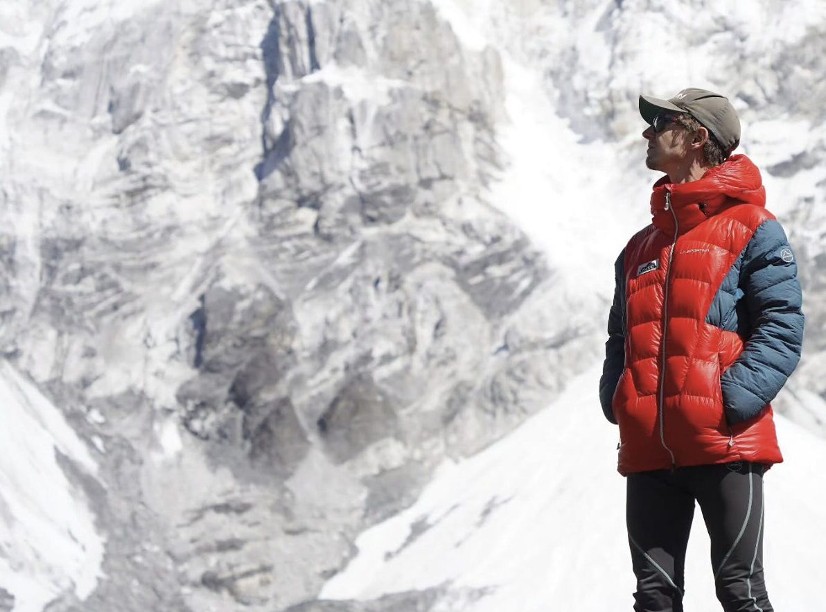A man wearing a red and blue down jacket, black leggings, sunglasses, and a cap stands in front of a towering, snow-covered mountain face, looking upward. The rugged alpine backdrop suggests a high-altitude location, likely in the Himalayas.