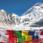 Colorful Tibetan prayer flags flutter in the foreground with a stunning view of snow-covered peaks in the Himalayas, including Mount Everest and its surrounding glaciers, under a clear blue sky.