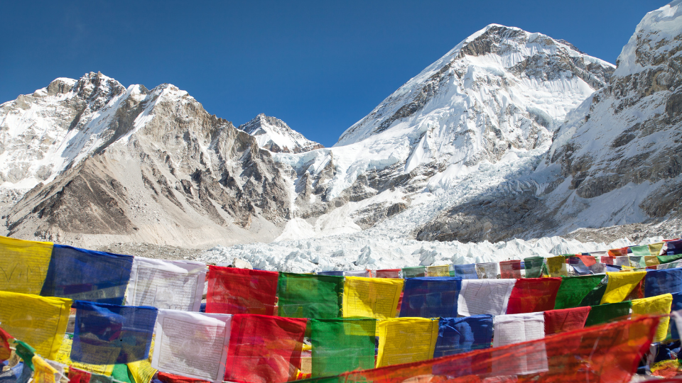 Colorful Tibetan prayer flags flutter in the foreground with a stunning view of snow-covered peaks in the Himalayas, including Mount Everest and its surrounding glaciers, under a clear blue sky.