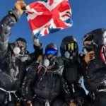 Five climbers wearing black down suits and oxygen masks stand on the summit of Mount Everest, proudly holding up a British flag against a clear blue sky.
