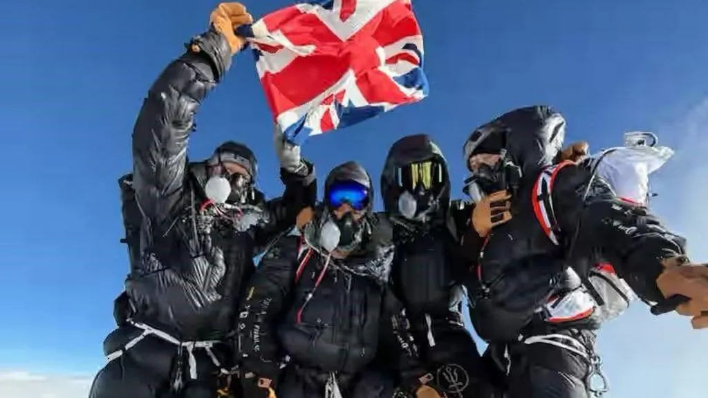 Five climbers wearing black down suits and oxygen masks stand on the summit of Mount Everest, proudly holding up a British flag against a clear blue sky.