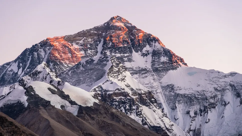 Mount Everest at sunrise, viewed from the Tibetan side, with the summit and upper ridges bathed in a warm orange glow while the lower slopes remain in shadow, showcasing the mountain’s rugged, snow-covered terrain under a clear sky.