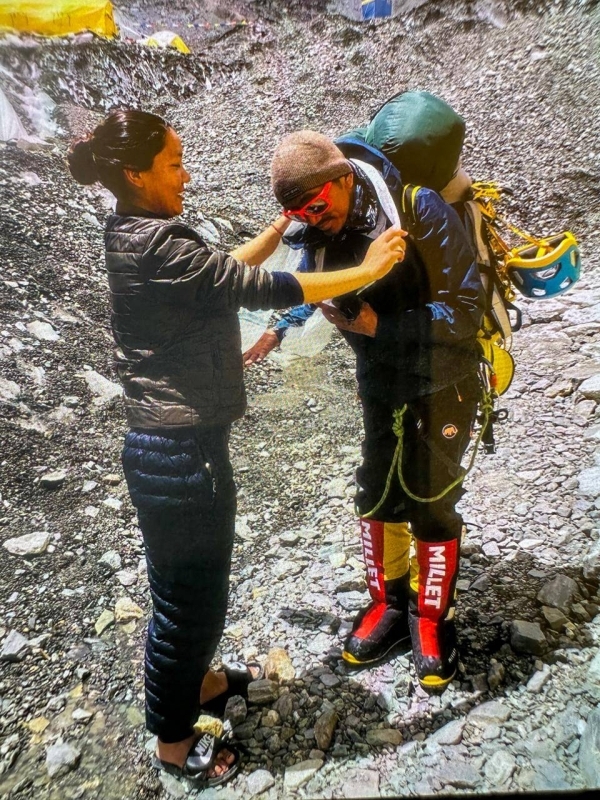 Two people in a rocky mountain setting, one adjusting the other's gear. The individual being assisted wears a backpack, climbing equipment, and red "Millet" boots, while the other is dressed in a dark puffer jacket and sandals. Tents are visible in the background.
