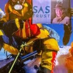 A mountaineer in colorful gear holding a sign reading "SAS TRUST" with a photo of an older man in a hat, set against a snowy mountain backdrop.