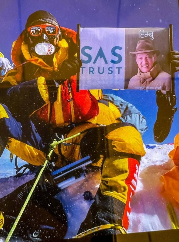 A mountaineer in colorful gear holding a sign reading "SAS TRUST" with a photo of an older man in a hat, set against a snowy mountain backdrop.