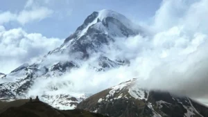 Snow-covered Mount Kazbek rises dramatically through a blanket of clouds, with the silhouette of the Gergeti Trinity Church visible on a hill in the foreground.