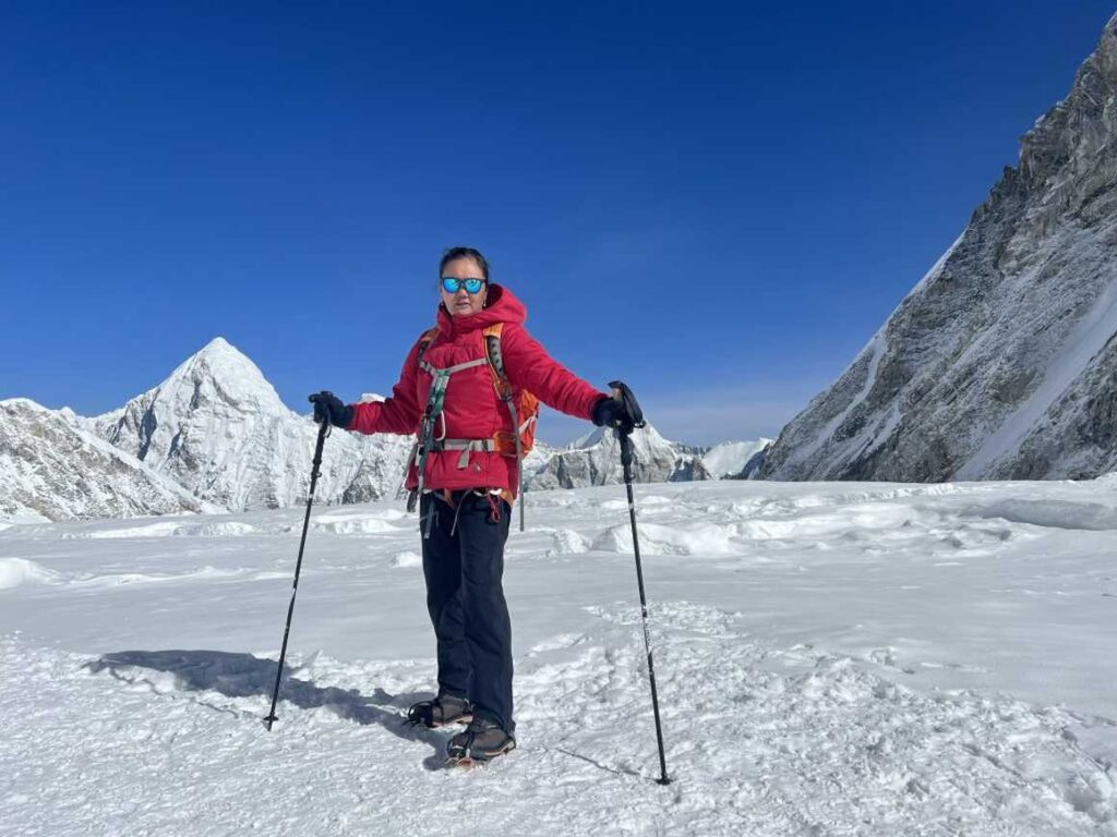 A climber wearing a red jacket, sunglasses, and a backpack stands confidently on a snowy mountain plateau, holding trekking poles. Jagged snow-covered peaks rise in the background under a clear blue sky.