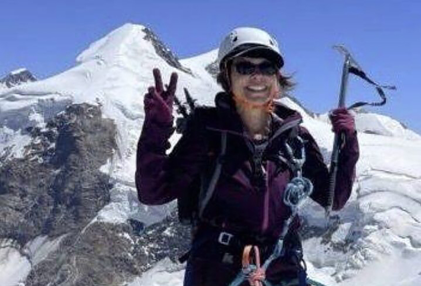 A smiling female mountaineer poses on a snowy mountain ridge, wearing a helmet, sunglasses, gloves, and climbing gear. She holds an ice axe in one hand and flashes a peace sign with the other, with snow-covered peaks and clear blue skies in the background