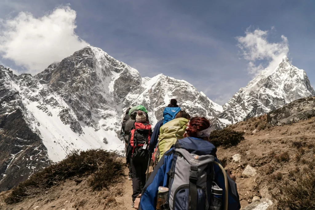 Group of trekkers ascending a mountain trail with snow-covered peaks in the Himalayas in the background, under clear blue skies