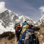 Group of trekkers ascending a mountain trail with snow-covered peaks in the Himalayas in the background, under clear blue skies