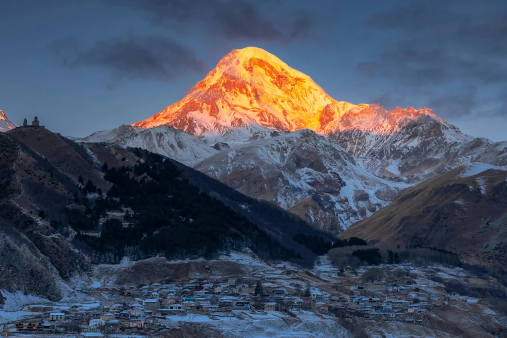 Sunrise illuminating the snow-covered peak of Mount Kazbek, with the village of Stepantsminda and Gergeti Trinity Church in the foreground, in the Caucasus Mountains of Georgi