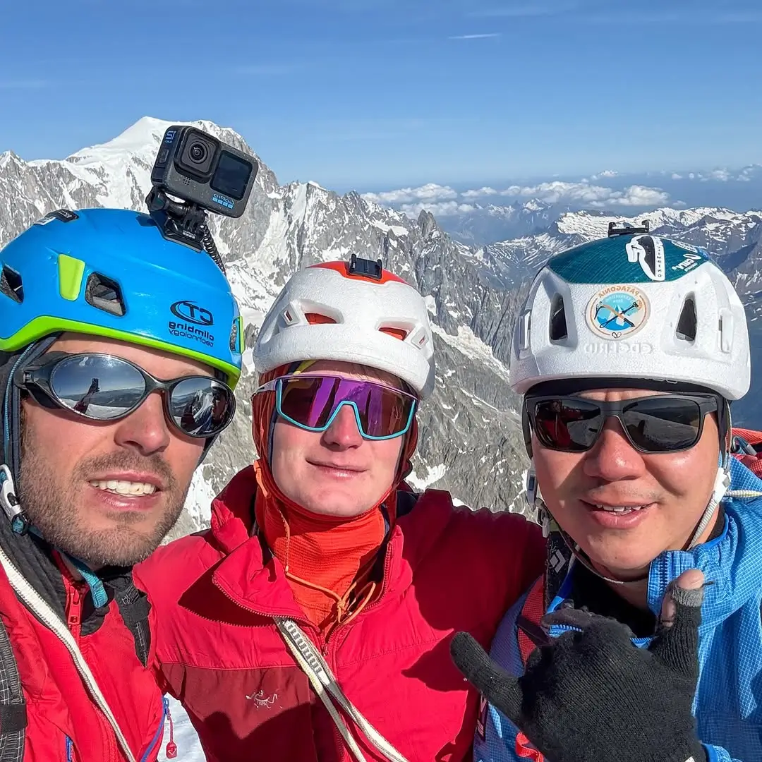 Three climbers wearing helmets and sunglasses smile for a summit selfie with snow-covered alpine peaks in the background after successfully completing the Grandes Jorasses ascent