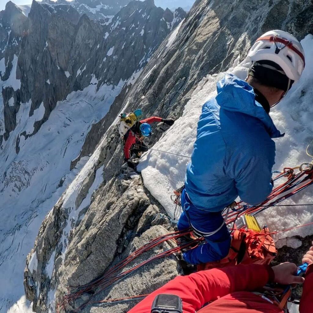 "Climbers secured with ropes on a steep, icy rock face of the Grandes Jorasses, carefully navigating a mixed climbing section with snow, ice, and rock exposure."