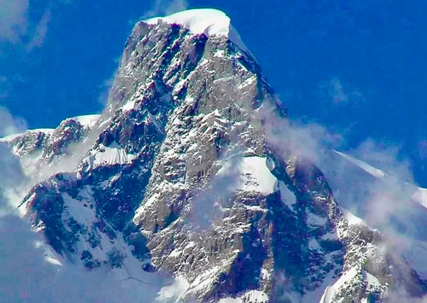 **Alt text:** Snow-covered Southeast Pillar of Ultar Sar towering sharply against a clear blue sky, with rugged rock faces and light clouds swirling around the flanks.
