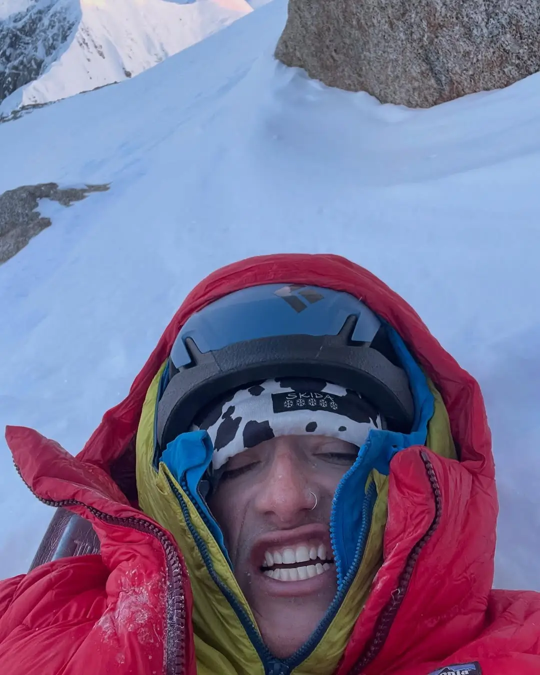 A close-up selfie of a climber bundled in a red and yellow down jacket, helmet, and headband, grimacing or smiling with eyes closed, surrounded by snow and high alpine terrain on Denali’s steep south face.